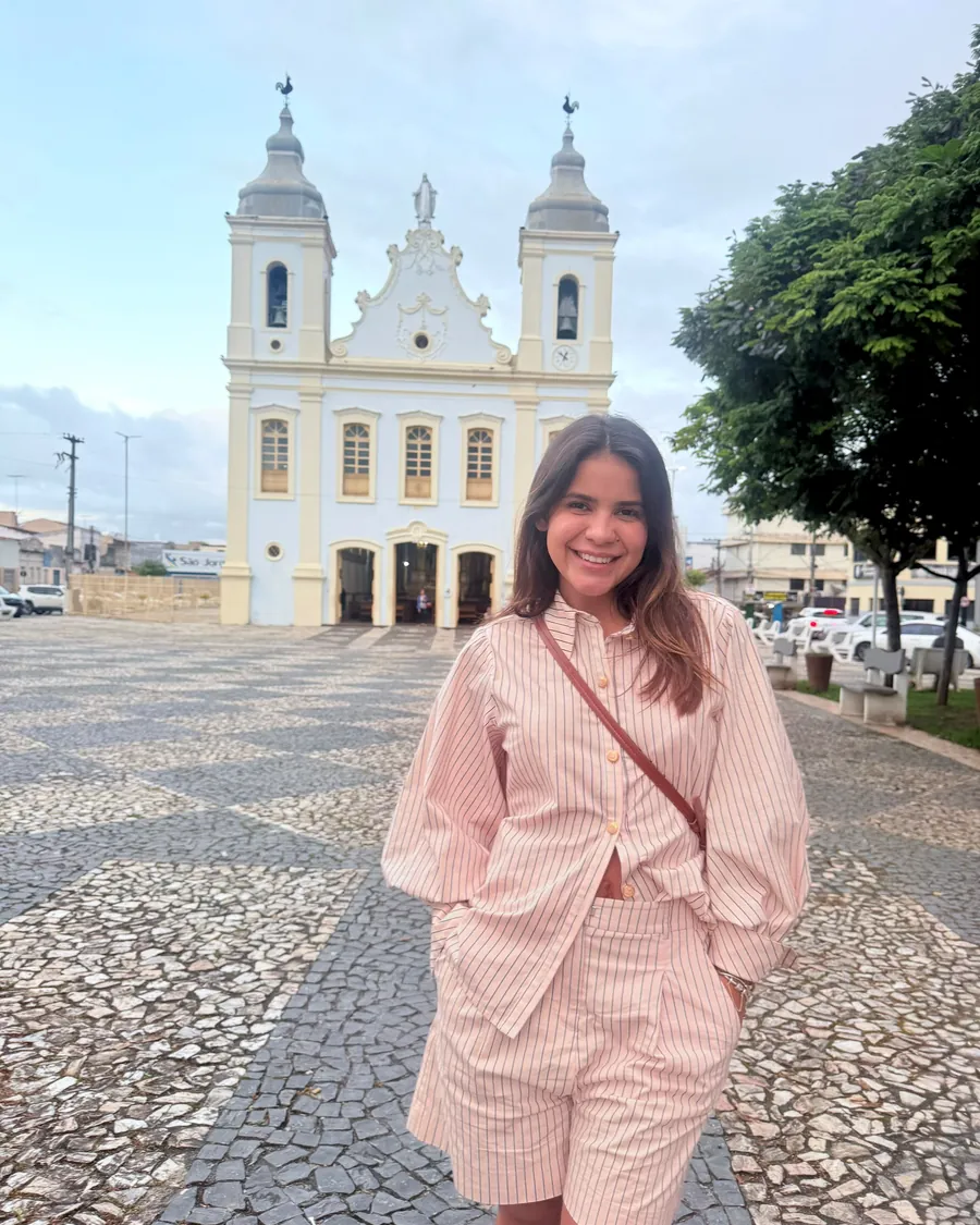 Gabriella Moraes sorrindo, de camisa listrada rosa, em uma praça com igreja ao fundo